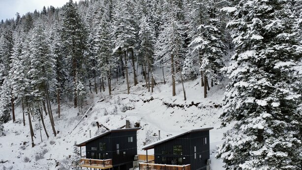 A modern cabin with a wooden deck and a small wooden structure nearby, surrounded by a snowy forest.