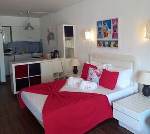 Quarto de hotel com cama, mesas de cabeceira, escrivaninha, cadeira, abajur, prateleira com garrafas, um quadro e uma área de cozinha.