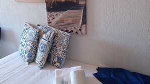 Ein ordentlich bezogenes Bett mit weißer Decke, zwei gemusterten Kissen, einem gefalteten weißen Handtuch und einer blauen Decke.