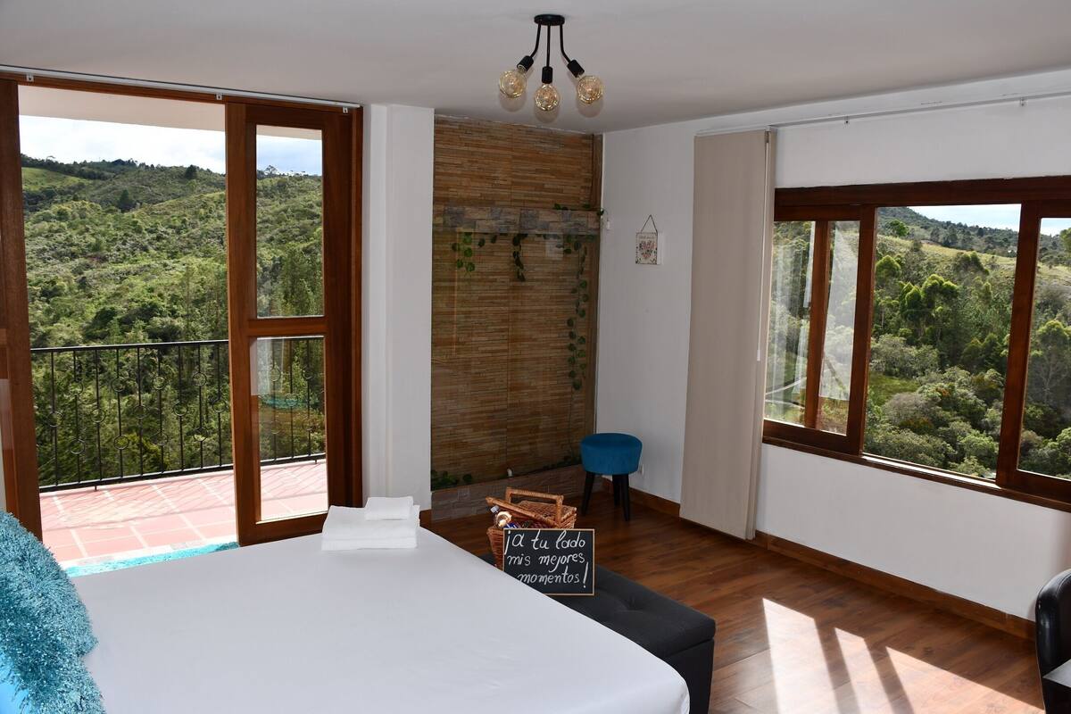 A bedroom with a bed, a small chair, a wooden floor, a balcony with a view of greenery, and a wall with a wooden panel and a hanging light fixture.