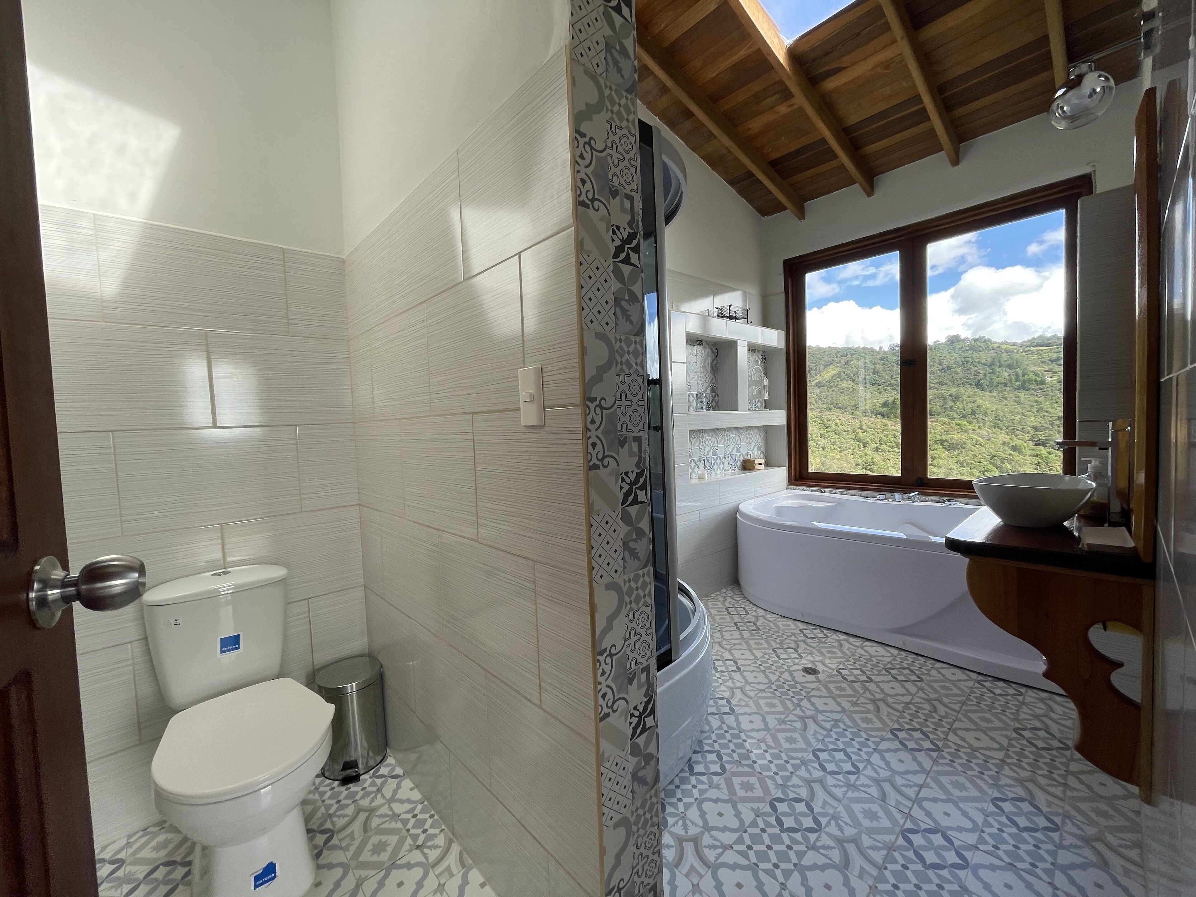 Un baño con inodoro, lavamanos y bañera. Se ve un ventanal con vistas a un paisaje montañoso.