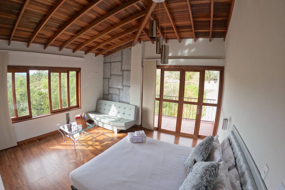 A spacious bedroom with a large bed, a seating area, and a view of greenery through the windows.