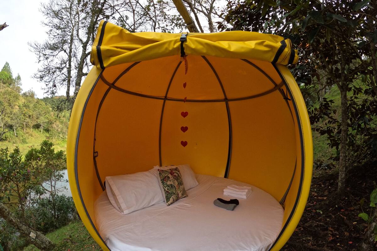 A round, yellow hanging bed with a white mattress and pillows, set in a natural outdoor environment with trees and a body of water in the background.