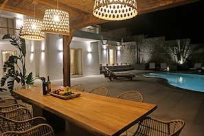 An outdoor dining area with a wooden table and wicker chairs, a pool, and a stone wall in the background.