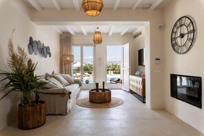 A spacious living room with a sofa, a coffee table, and a view of a pool and outdoor seating area. - Island Times Villa by Unique Villas Collection (Mykonos)