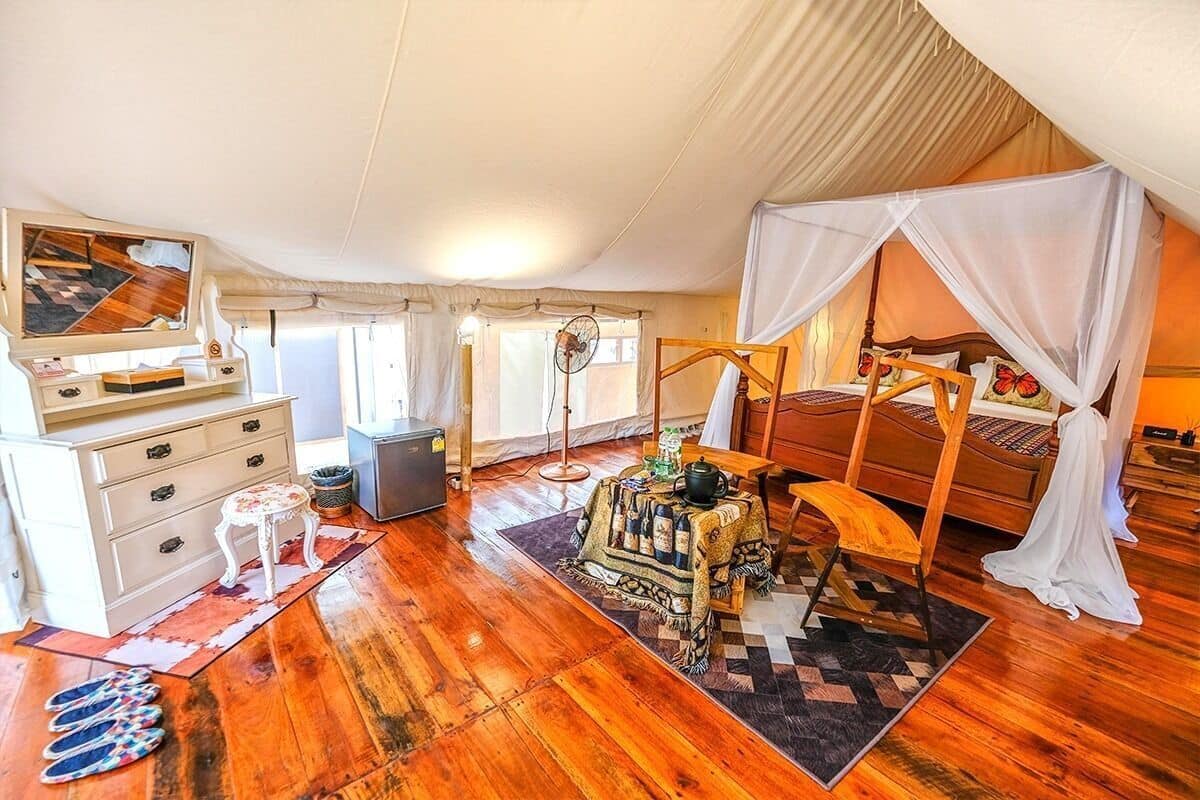 A tent-style room with a bed, a small table with a tablecloth, a chair, a dresser, a mirror, and a fan.