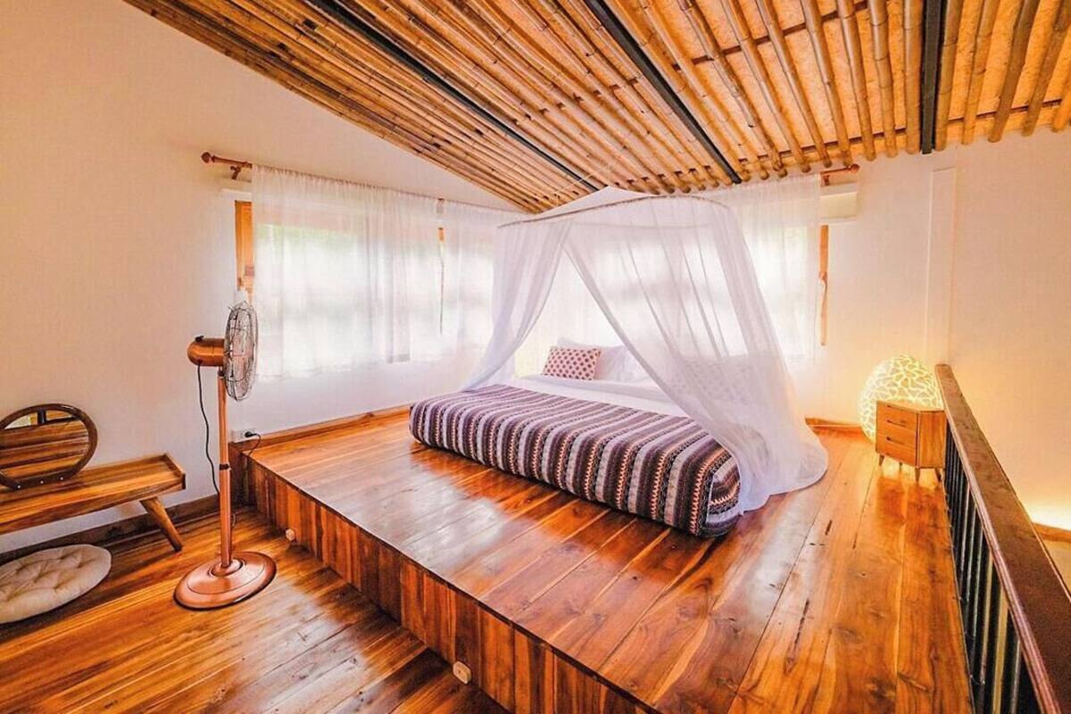 A room with a bed under a canopy, a standing fan, a bench, and a nightstand with a lamp.
