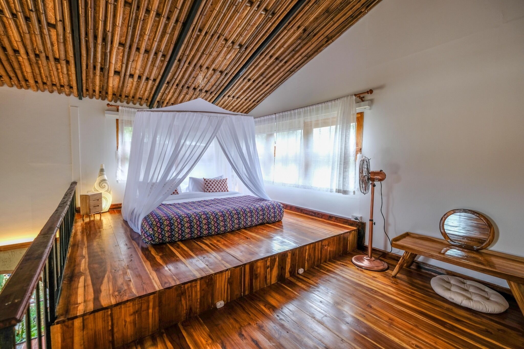 Un dormitorio con una cama plataforma de madera, dosel blanco, lámpara de noche y un espejo redondo sobre un estante de madera.