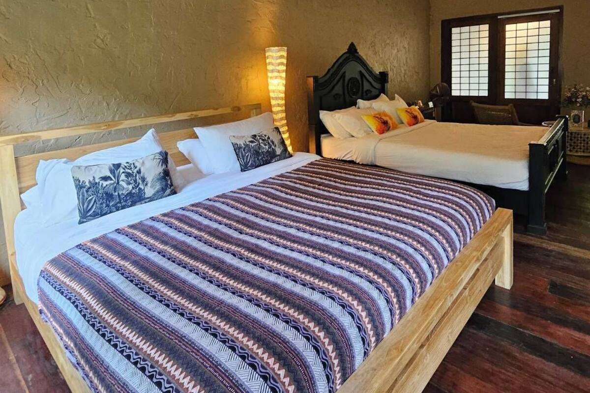 A bedroom with a large bed, a smaller bed, a nightstand, a lamp, and a window with traditional shutters.