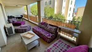 A balcony with a sofa, a small table, and a chair. There is a railing and a view of buildings and trees.