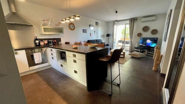 A modern kitchen with a central island, a dining area with a bar stool, and a living room with a television.
