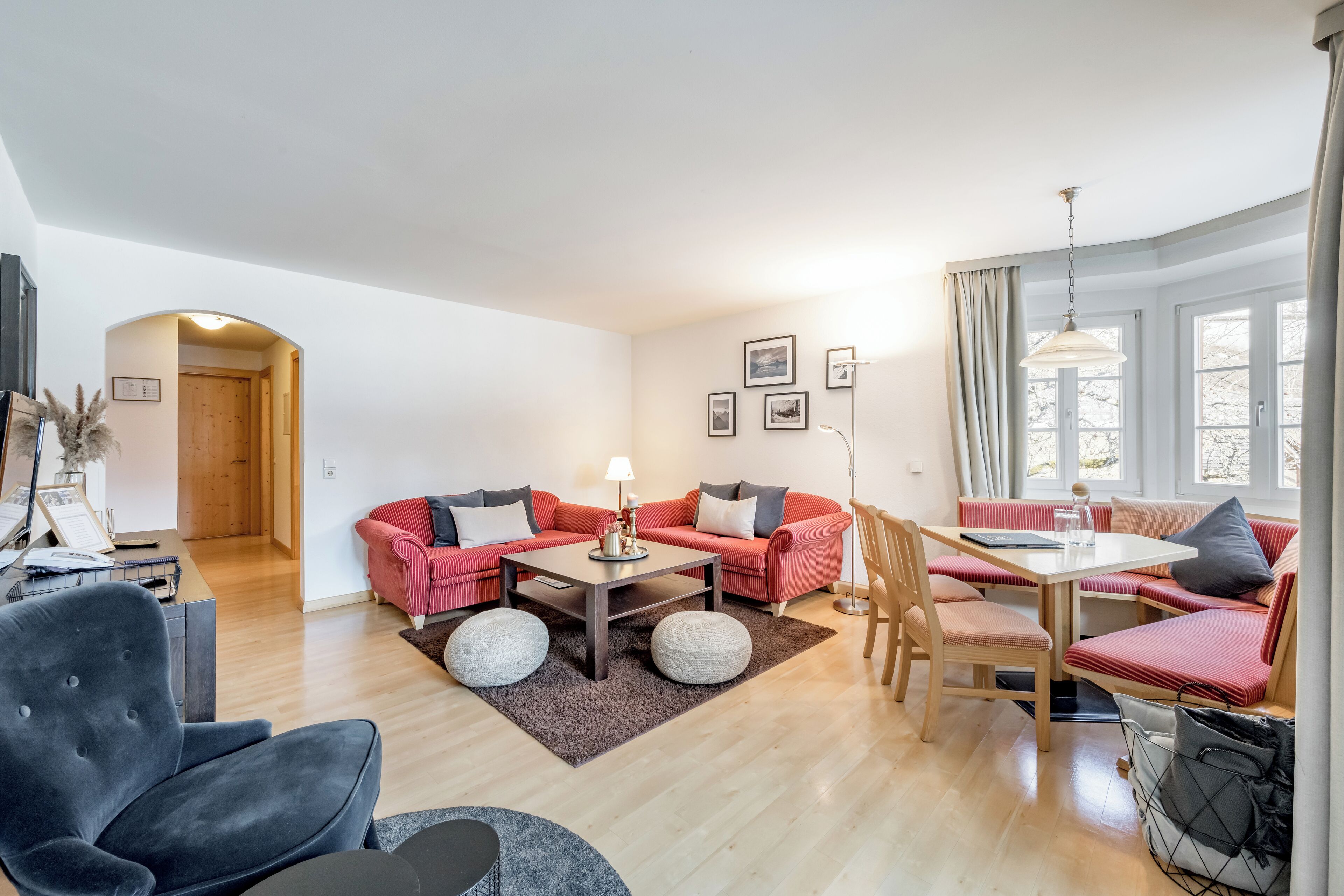 Un salon moderne avec un ensemble de canapés rouges, un fauteuil noir, une table à manger en bois et une grande fenêtre avec des rideaux.