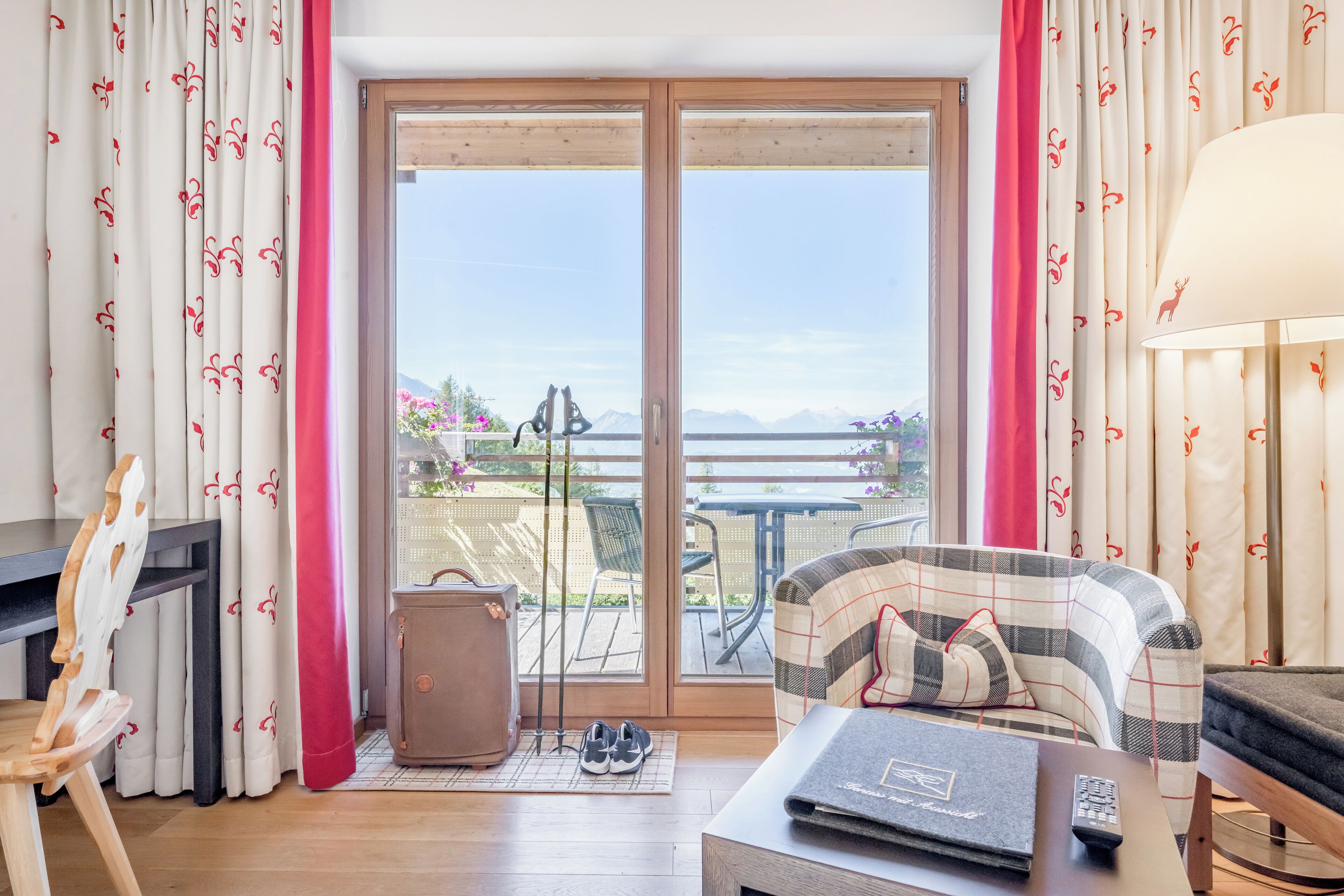Una habitación con piso de madera, un sofá, una mesa con un control remoto, una maleta y vista a un balcón con mobiliario exterior y montañas al fondo.
