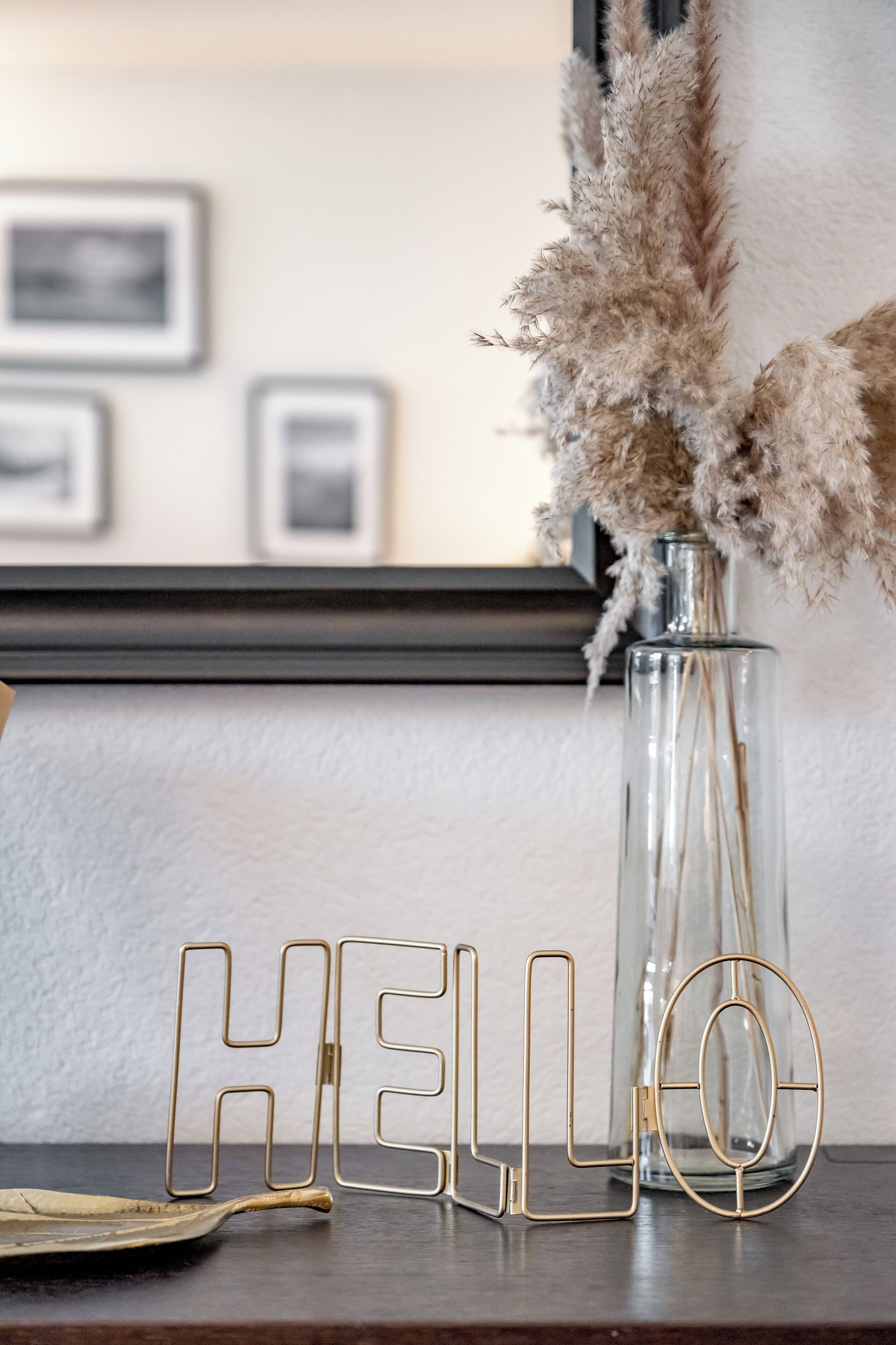 Eine klare Glasvase mit Pampasgras, ein goldener Schriftzug "HELLO" und ein goldener Tablett auf einer Holztischfläche.