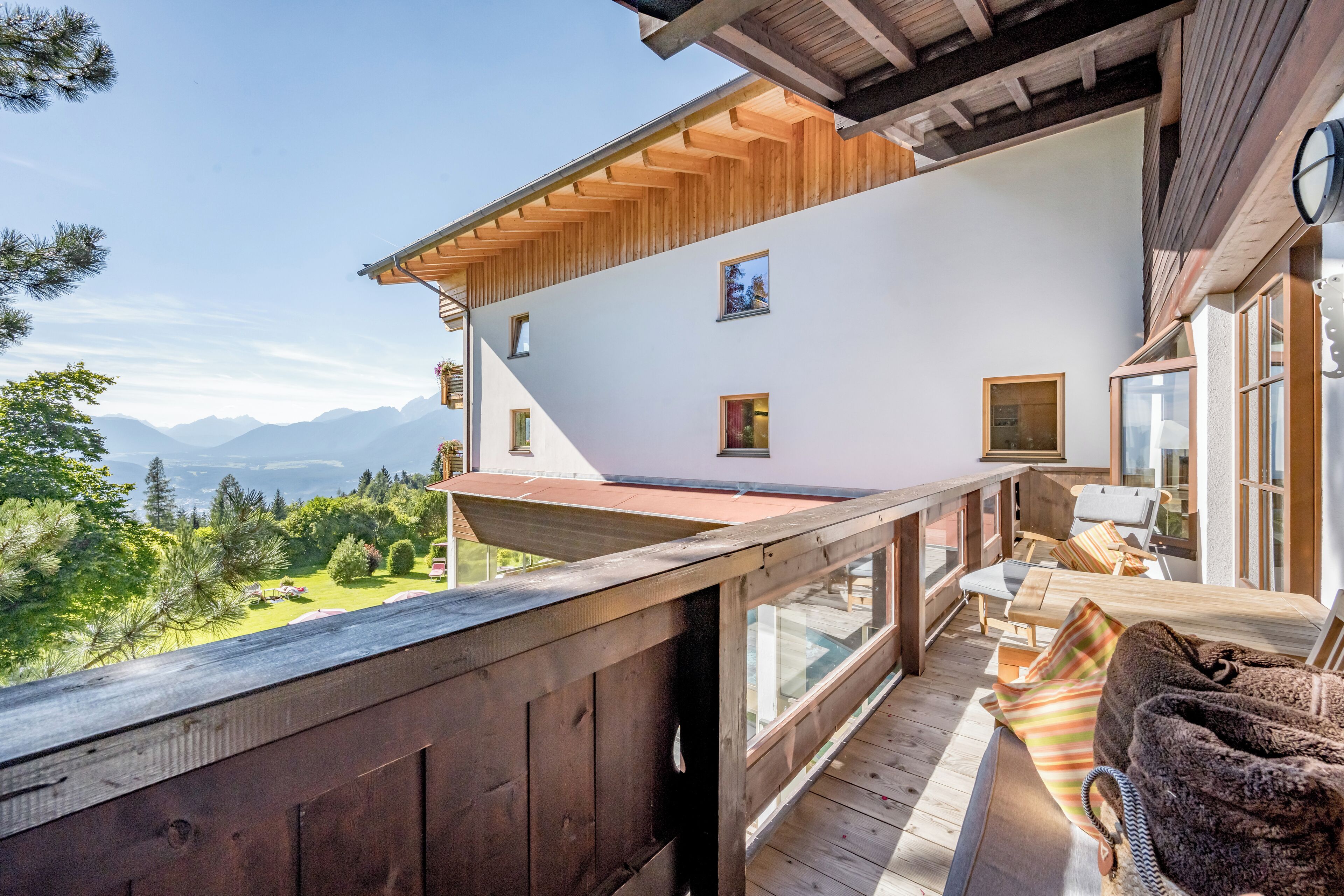 Una terraza de madera con vistas a montañas, un espacio de descanso cubierto y un amplio ventanal que domina el paisaje.