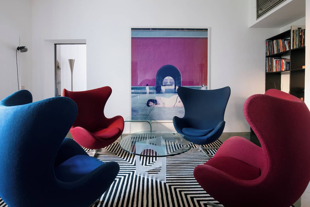 A modern room with a glass coffee table, red and blue chairs, a black and white patterned rug, and a large painting on the wall.