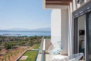 A balcony with a view of the sea and mountains, white chairs, and a glass door.