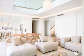 A modern living room with a high ceiling, a dining area with wooden furniture, and a neutral color palette.