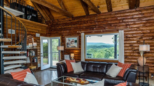 L’intérieur d’une cabane en rondins, avec un canapé en cuir, une table à manger sur laquelle est disposé un plateau de fruits, un escalier et une grande fenêtre donnant sur l’extérieur.