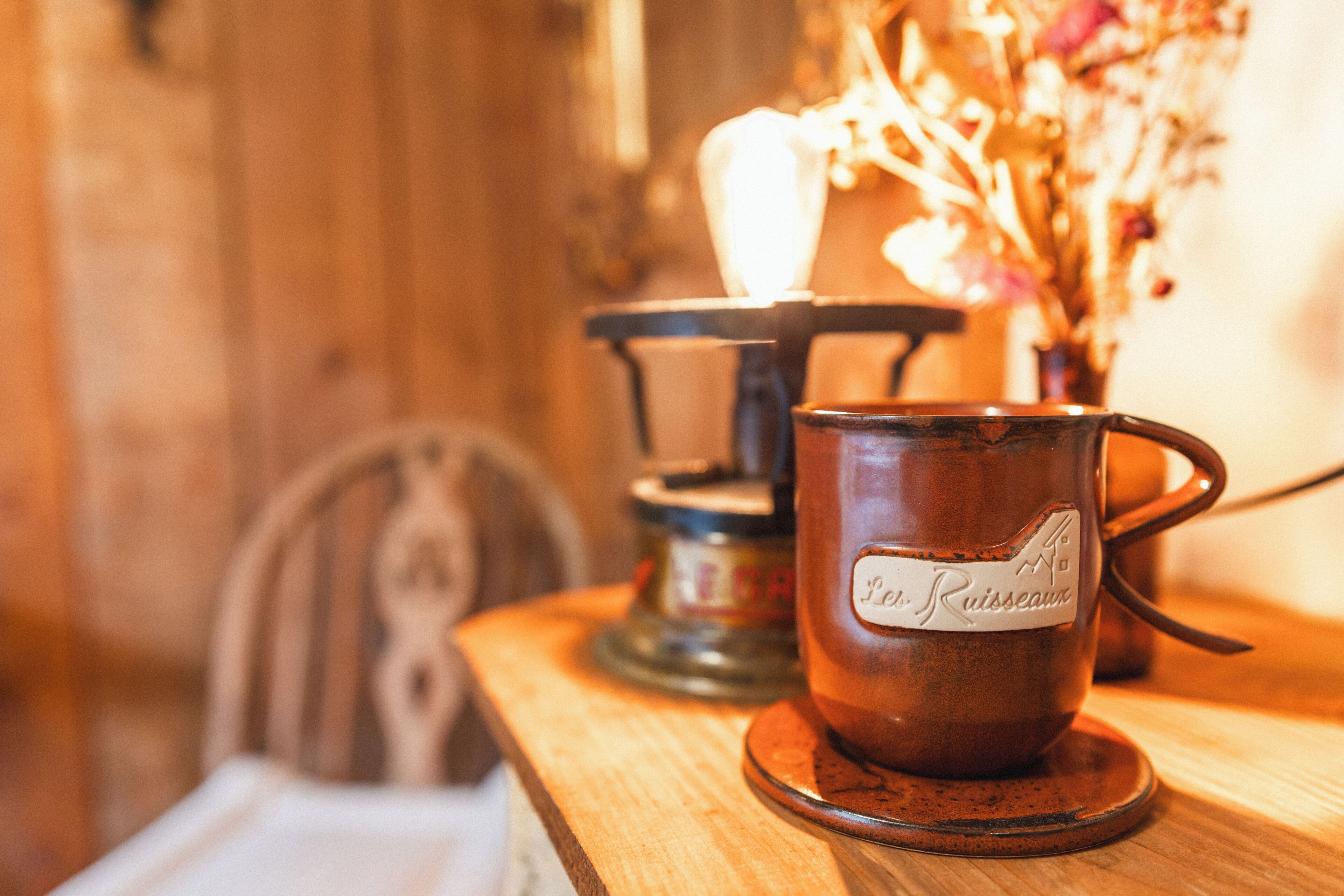 Una mesa de madera con un candelabro, una taza con etiqueta y un florero con flores secas.