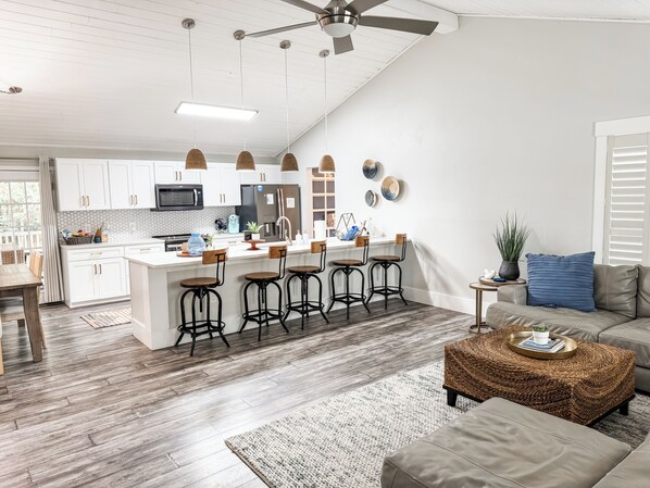 Una cocina moderna con armarios blancos, una isla central con taburetes, un ventilador de techo y un comedor con mesa y sillas de madera.