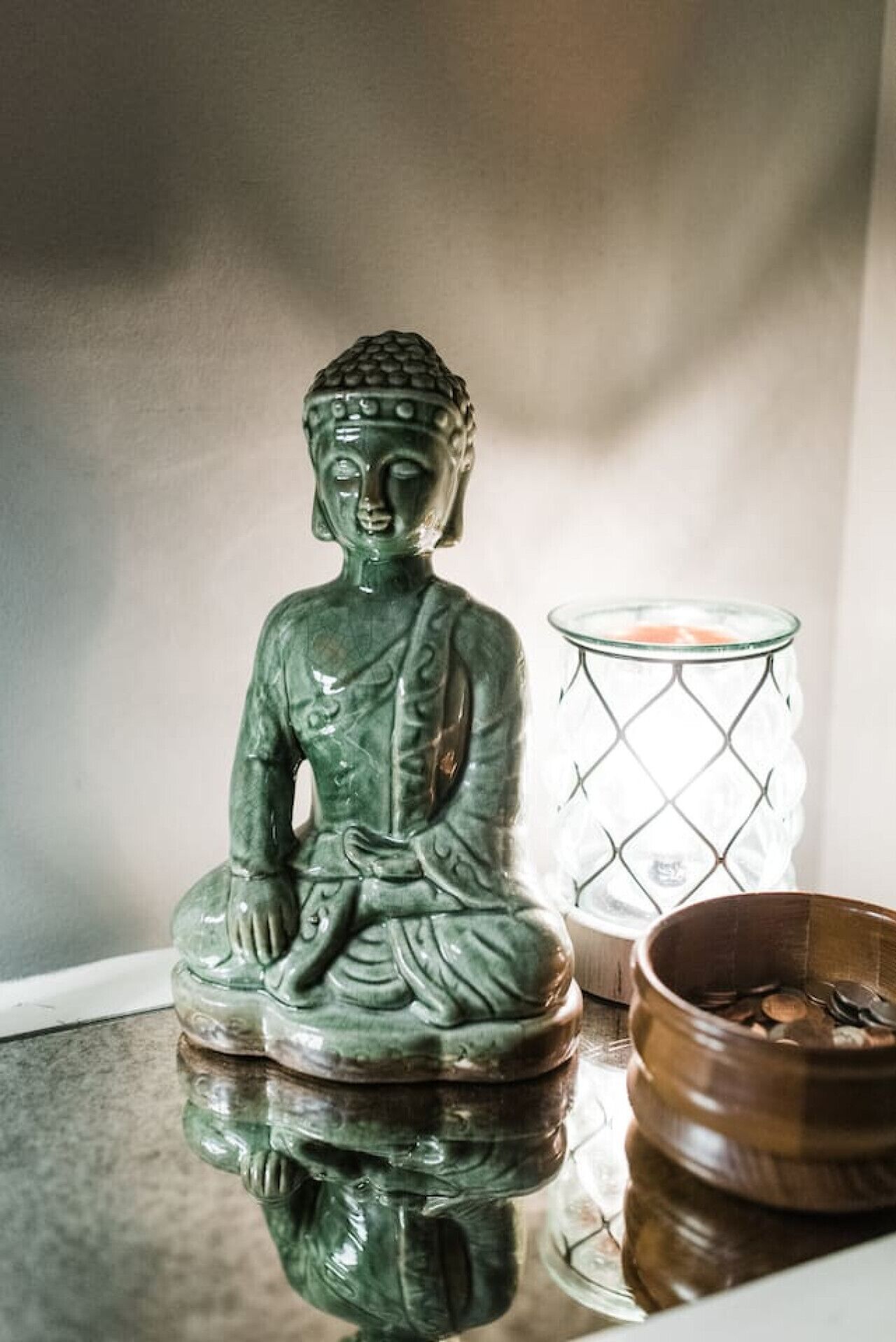 Une statue de Bouddha verte posée sur une surface réfléchissante, un porte-bougie en verre avec une bougie, et un bol en bois contenant des pièces de monnaie.