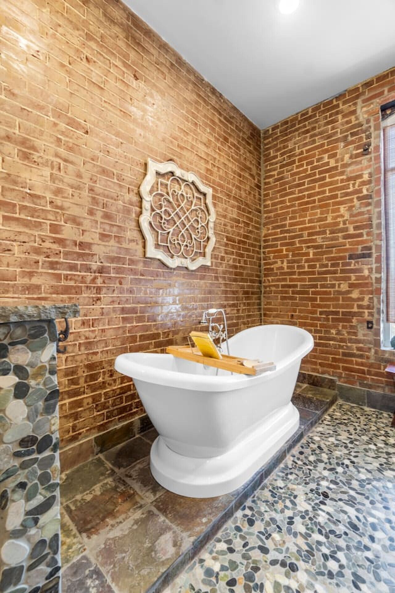 A bathroom with a white freestanding bathtub, a stone mosaic floor, and a brick wall with a decorative metal piece.