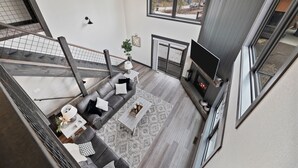 A modern living room with a grey sofa, coffee table, and fireplace. A large window offers a view of the outdoors. - Summit Sunset, pet friendly! Hot Tub, Pool access, paved roads, luxury finishes (Lead)