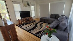 A living room with a grey sofa, a flat-screen TV on a wooden stand, a patterned rug, and a potted plant. - Recovery On Discovery Holiday House (Agnes Water)