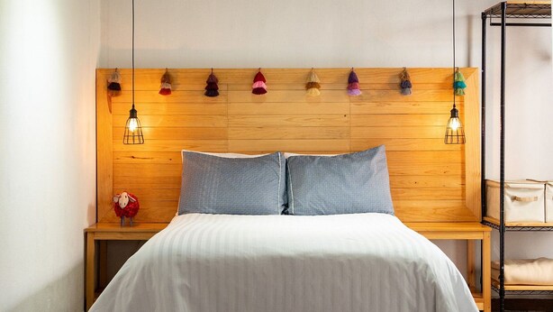 Een slaapkamer met een houten hoofdbord, een wit sprei, twee kussens, een nachtkastje met een lamp en een klein tafeltje met een handtas.