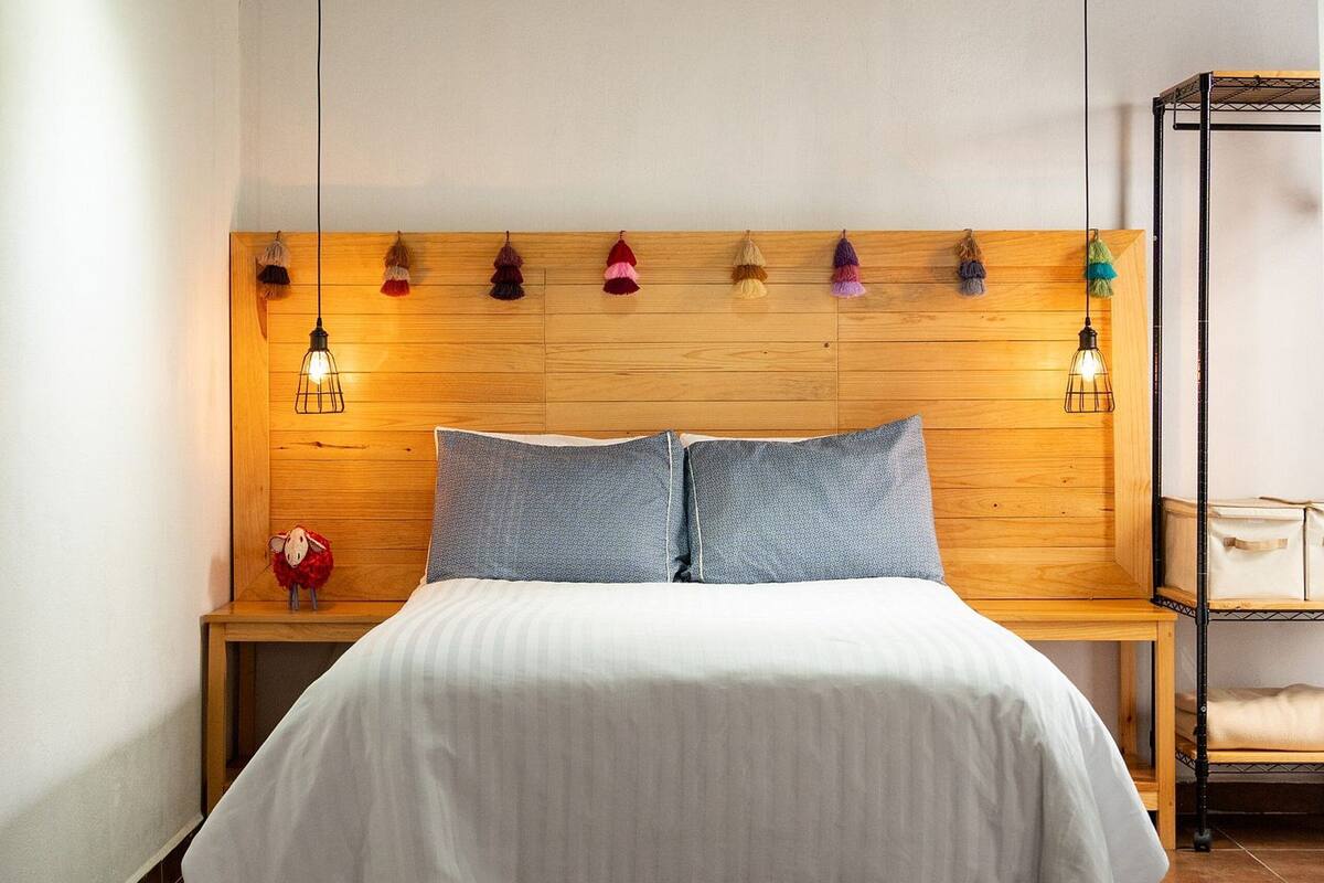 Ein Schlafzimmer mit einem hölzernen Kopfteil, einer weißen Bettdecke, zwei Kissen, einem Nachttisch mit einer Lampe und einem kleinen Tisch mit einer Handtasche.