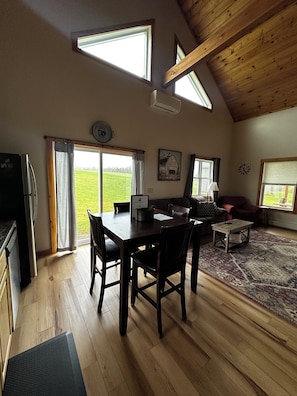 Une pièce spacieuse avec un plafond en bois, une table à manger avec des chaises, un canapé, une table basse et une vue sur un champ verdoyant grâce à de grandes fenêtres.