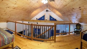 Quarto amplo com pé-direito alto, vigas de madeira e teto inclinado, com duas camas e roupa de cama colorida e um corrimão de madeira no centro.