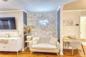 A modern living room with a brick wall, a flat-screen TV, a sofa, a coffee table, and a desk with a chair. - POOL with Heating option! 6-seater Golf Cart &GameRoom! Walk to Beach &Downtown! (Long Beach)