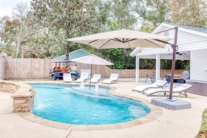 A swimming pool with a curved edge, surrounded by lounge chairs and umbrellas, with a wooden fence and trees in the background.