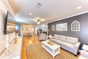 A spacious living room with a grey sofa, a coffee table, and a patterned rug. There is a dining area with a white table and chairs, and a kitchen in the background. - POOL with Heating option! 6-seater Golf Cart &GameRoom! Walk to Beach &Downtown! (Long Beach)
