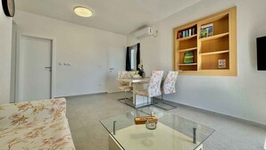 Una sala de estar moderna con un sofá, una mesa de centro de vidrio, un comedor con sillas, una estantería y una puerta.