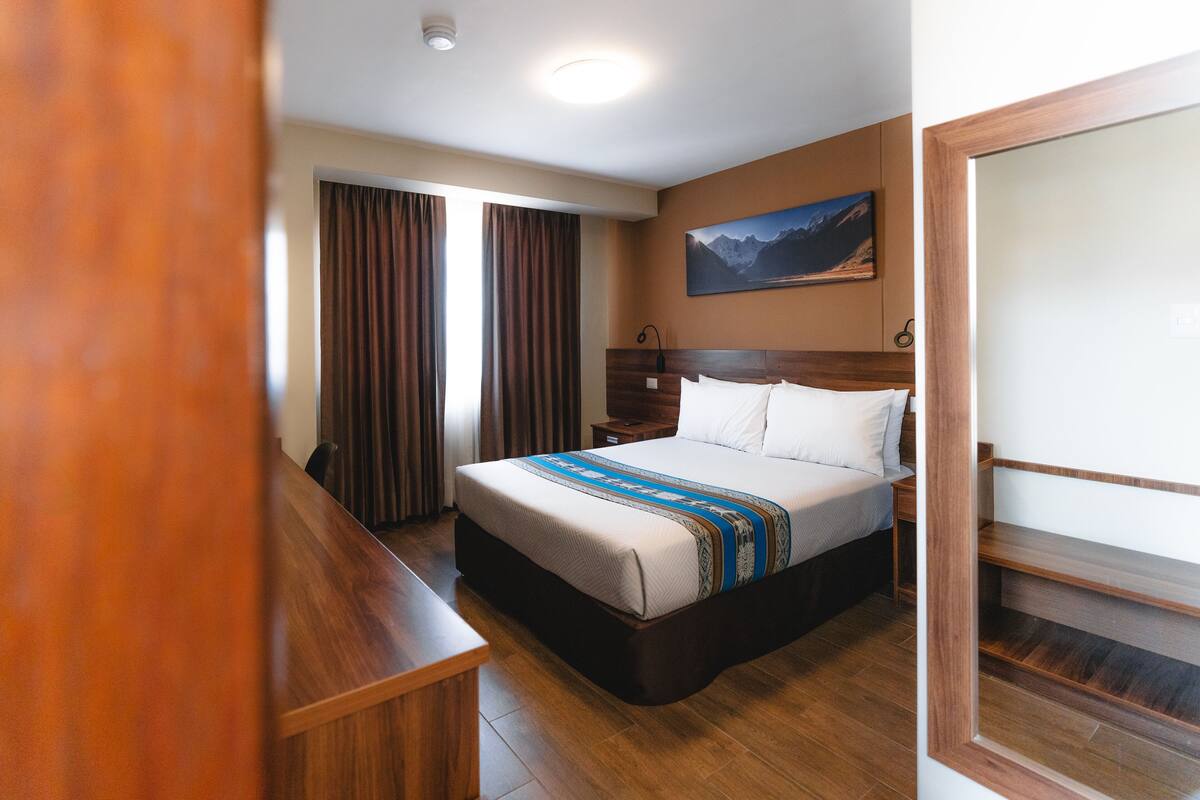 A hotel room with a bed, wooden floor, curtains, a painting on the wall, and a desk.