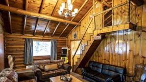 A cozy cabin interior with a wooden ceiling, a staircase, a sofa, a coffee table, and a fireplace.