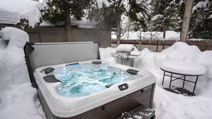 A hot tub in a snowy backyard with a grill and a covered table.