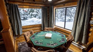 A wooden cabin with a round table and chairs, a window with curtains, and a view of snow-covered trees.