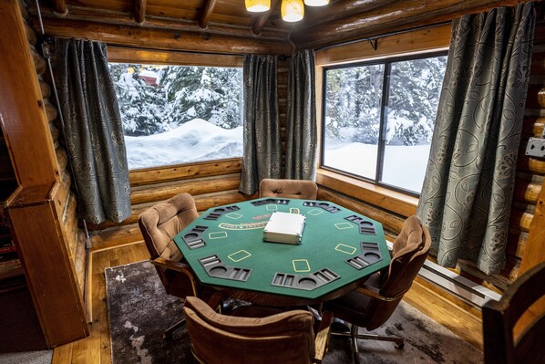 A wooden cabin with a round table and chairs, a window with curtains, and a view of snow-covered trees.