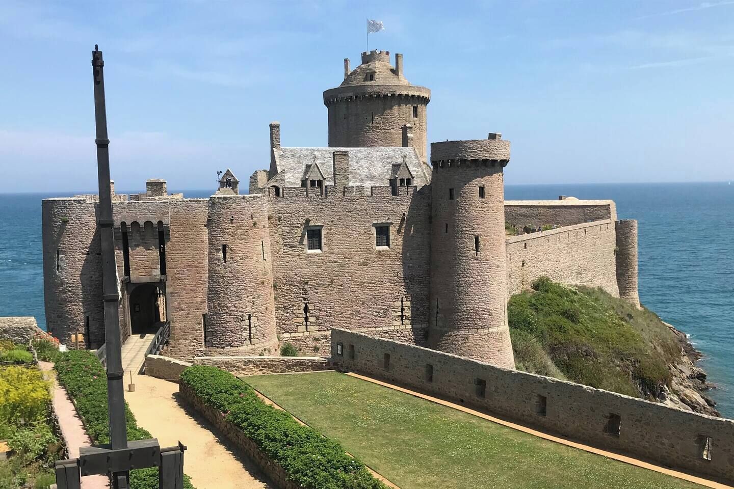 A historic stone castle with a prominent tower, a fortified wall, and a view of the sea.