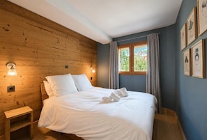 A bedroom with a wooden wall, a bed with white linens, a nightstand, and a window with curtains.