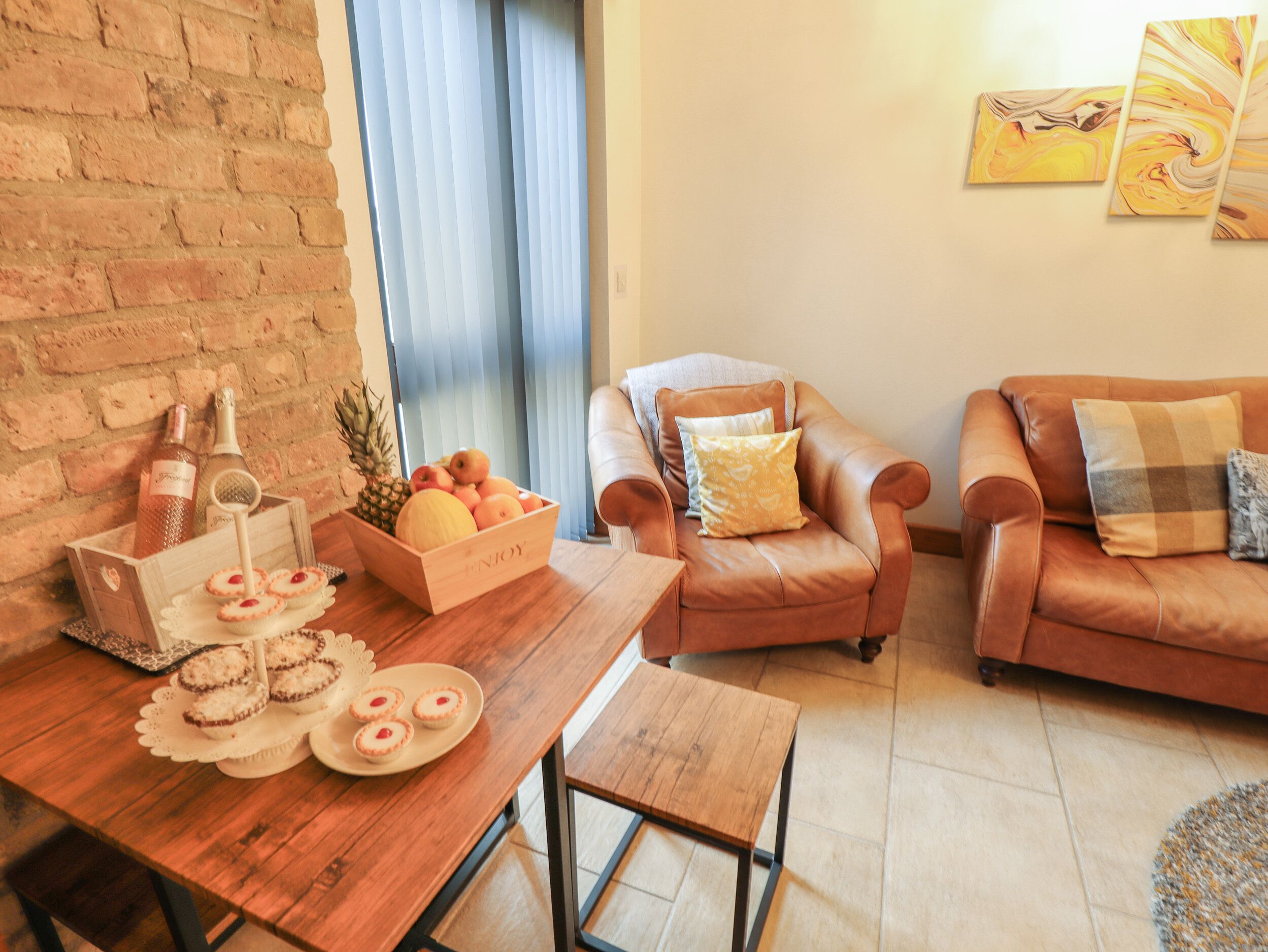 A cozy living room with a wooden table, a leather armchair, and a sofa. There is a fruit basket on the table and a bottle of wine.