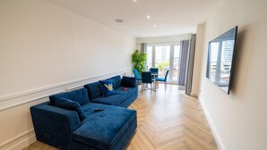 A modern living room with a large blue sectional sofa, a dining area with a table and chairs, and a large window offering a view of the city. - Stunning Penthouse - Birmingham City Centre (Birmingham)