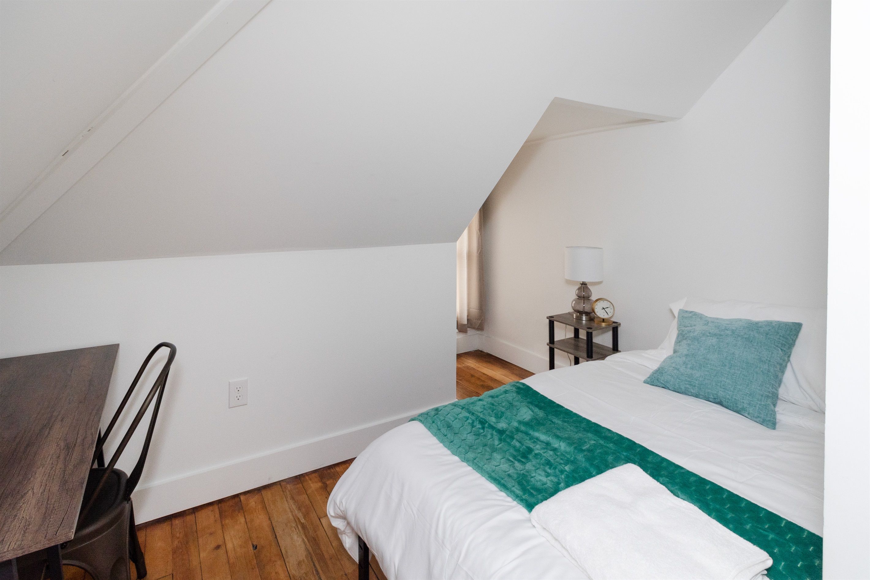 A bedroom with a sloped ceiling, a bed with a green and white bedspread, a nightstand with a lamp, and a chair.