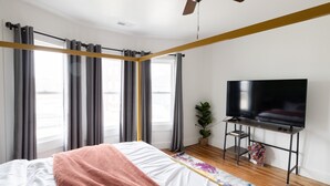 Une chambre à coucher avec un lit, un téléviseur posé sur un meuble, un ventilateur de plafond et de grandes fenêtres avec des rideaux.
