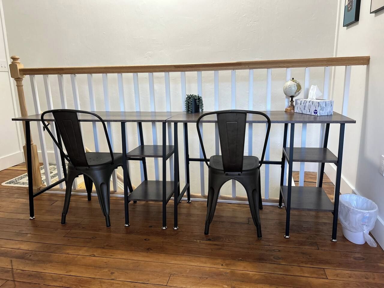 A dining area with a wooden railing, two black chairs, a small table, and a trash bin.