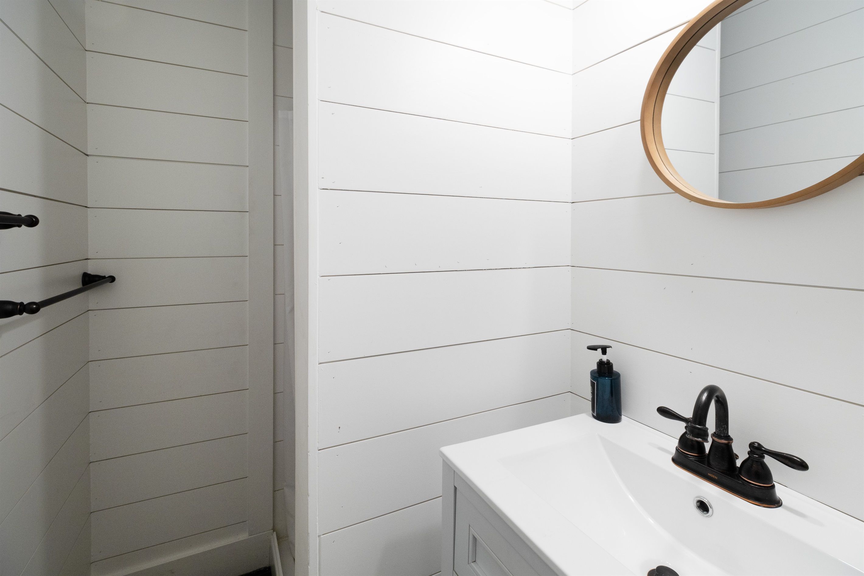 A bathroom with white wooden paneling, a white sink with a black faucet, a round mirror, and a black soap dispenser.
